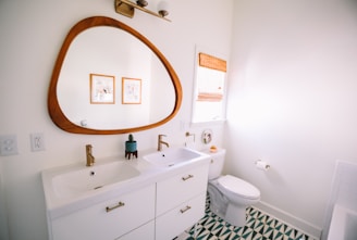 A modern bathroom with a large wooden-framed mirror above a dual sink vanity. The sinks have gold faucets, and the vanity is white with golden handles. A toilet is situated next to the sink, beneath a window covered with a woven blind. The floor features a green and white geometric pattern. Two framed pictures hang on the wall.