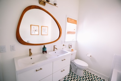 A modern bathroom with a large wooden-framed mirror above a dual sink vanity. The sinks have gold faucets, and the vanity is white with golden handles. A toilet is situated next to the sink, beneath a window covered with a woven blind. The floor features a green and white geometric pattern. Two framed pictures hang on the wall.