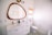A modern bathroom with a large wooden-framed mirror above a dual sink vanity. The sinks have gold faucets, and the vanity is white with golden handles. A toilet is situated next to the sink, beneath a window covered with a woven blind. The floor features a green and white geometric pattern. Two framed pictures hang on the wall.