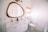 A modern bathroom with a large wooden-framed mirror above a dual sink vanity. The sinks have gold faucets, and the vanity is white with golden handles. A toilet is situated next to the sink, beneath a window covered with a woven blind. The floor features a green and white geometric pattern. Two framed pictures hang on the wall.