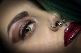 A close-up of a person's face highlighting the eye, nose, and lips. The eye is accentuated with glittery eyeshadow, bold eyeliner, and long lashes. There are multiple piercings on the nose, including a hoop and ball studs. The lips are painted with a dark, glossy red lipstick. The hair appears to be green and is visible at the top of the image.
