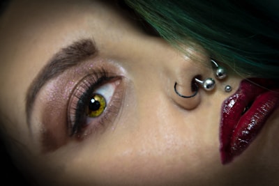 A close-up of a person's face highlighting the eye, nose, and lips. The eye is accentuated with glittery eyeshadow, bold eyeliner, and long lashes. There are multiple piercings on the nose, including a hoop and ball studs. The lips are painted with a dark, glossy red lipstick. The hair appears to be green and is visible at the top of the image.