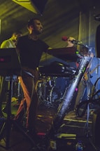 A band is performing on stage with a man singing into a red-colored microphone. He is accompanied by other musicians playing instruments. The setup includes electronic equipment and a drum kit in the background. Stage lighting creates a dramatic ambience with beams of light.