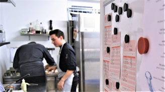 Two people in a professional kitchen are preparing food. One person is focused on cooking while the other seems to be observing. A whiteboard with lists and black magnets is visible in the foreground.