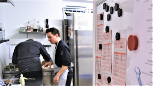 Two people in a professional kitchen are preparing food. One person is focused on cooking while the other seems to be observing. A whiteboard with lists and black magnets is visible in the foreground.