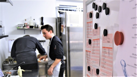 Two people in a professional kitchen are preparing food. One person is focused on cooking while the other seems to be observing. A whiteboard with lists and black magnets is visible in the foreground.