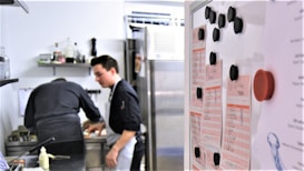 Two people in a professional kitchen are preparing food. One person is focused on cooking while the other seems to be observing. A whiteboard with lists and black magnets is visible in the foreground.
