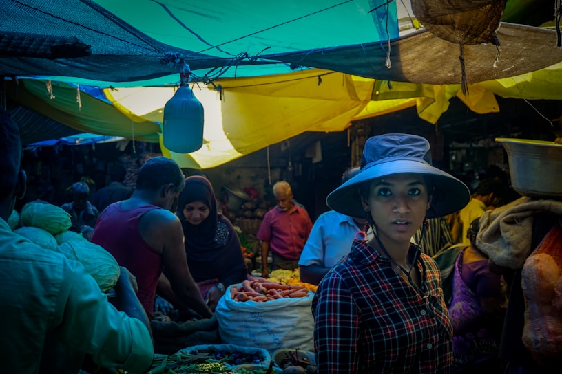 A bustling market scene with people engaging in trade. A woman wearing a wide-brimmed hat and a plaid shirt stands prominently in the foreground. The vibrant market stalls are filled with various vegetables, and the area is covered by colorful tarps creating a mix of light and shadow.