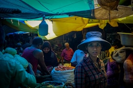 A bustling market scene with people engaging in trade. A woman wearing a wide-brimmed hat and a plaid shirt stands prominently in the foreground. The vibrant market stalls are filled with various vegetables, and the area is covered by colorful tarps creating a mix of light and shadow.