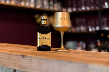A beautifully crafted bottle with a gold label sits alongside an elegant gold wine goblet on a wooden bar surface. The background is blurred, showcasing shelves with various glassware, creating a cozy and sophisticated bar atmosphere.