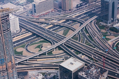 An aerial view of a complex multi-layered highway interchange surrounded by urban buildings. Multiple roads and overpasses create an intricate network of transportation infrastructure. The area is bustling with traffic, and nearby tall buildings suggest a bustling urban environment.