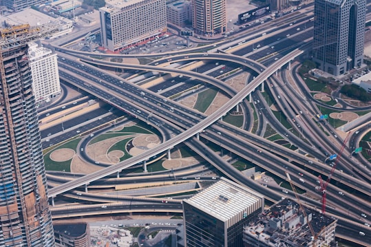 An aerial view of a complex multi-layered highway interchange surrounded by urban buildings. Multiple roads and overpasses create an intricate network of transportation infrastructure. The area is bustling with traffic, and nearby tall buildings suggest a bustling urban environment.