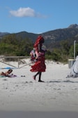 A person dressed in a vibrant red patterned outfit carries a bundle of colorful items, possibly bags or textiles, while walking on a sandy beach. In the background, there is a lush green landscape with mountains and a few scattered clouds in the blue sky. Another person is lying on the beach, partially covered with a towel.