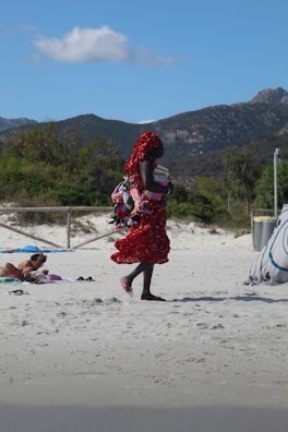 A person dressed in a vibrant red patterned outfit carries a bundle of colorful items, possibly bags or textiles, while walking on a sandy beach. In the background, there is a lush green landscape with mountains and a few scattered clouds in the blue sky. Another person is lying on the beach, partially covered with a towel.