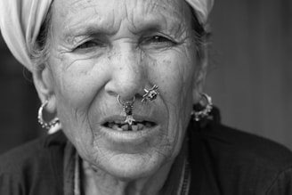 An older woman with distinct facial features and wearing traditional jewelry, including nose and ear rings. She has a headscarf and her skin shows visible signs of aging. Her expression is neutral, and the image is in black and white.