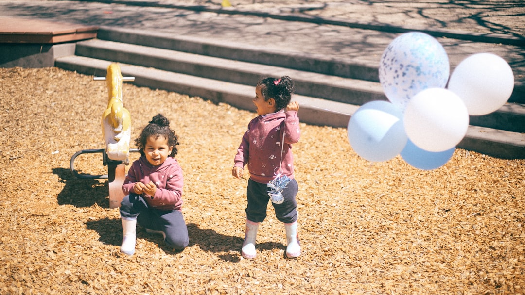 two twin girls playing on ground while holding white and blue balloons during daytime