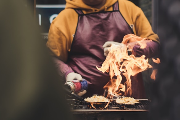 A person in a yellow hoodie and a dark apron is using a blow torch to cook dishes placed on a grill. Flames are visibly rising from the food, indicating high heat and a dynamic cooking process.