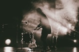 A person is energetically playing a drum on stage, surrounded by dramatic lighting and smoke effects. There is a microphone positioned near the drum set, and a guitar rests against the drum. The scene is cast in a monochrome, sepia tone.