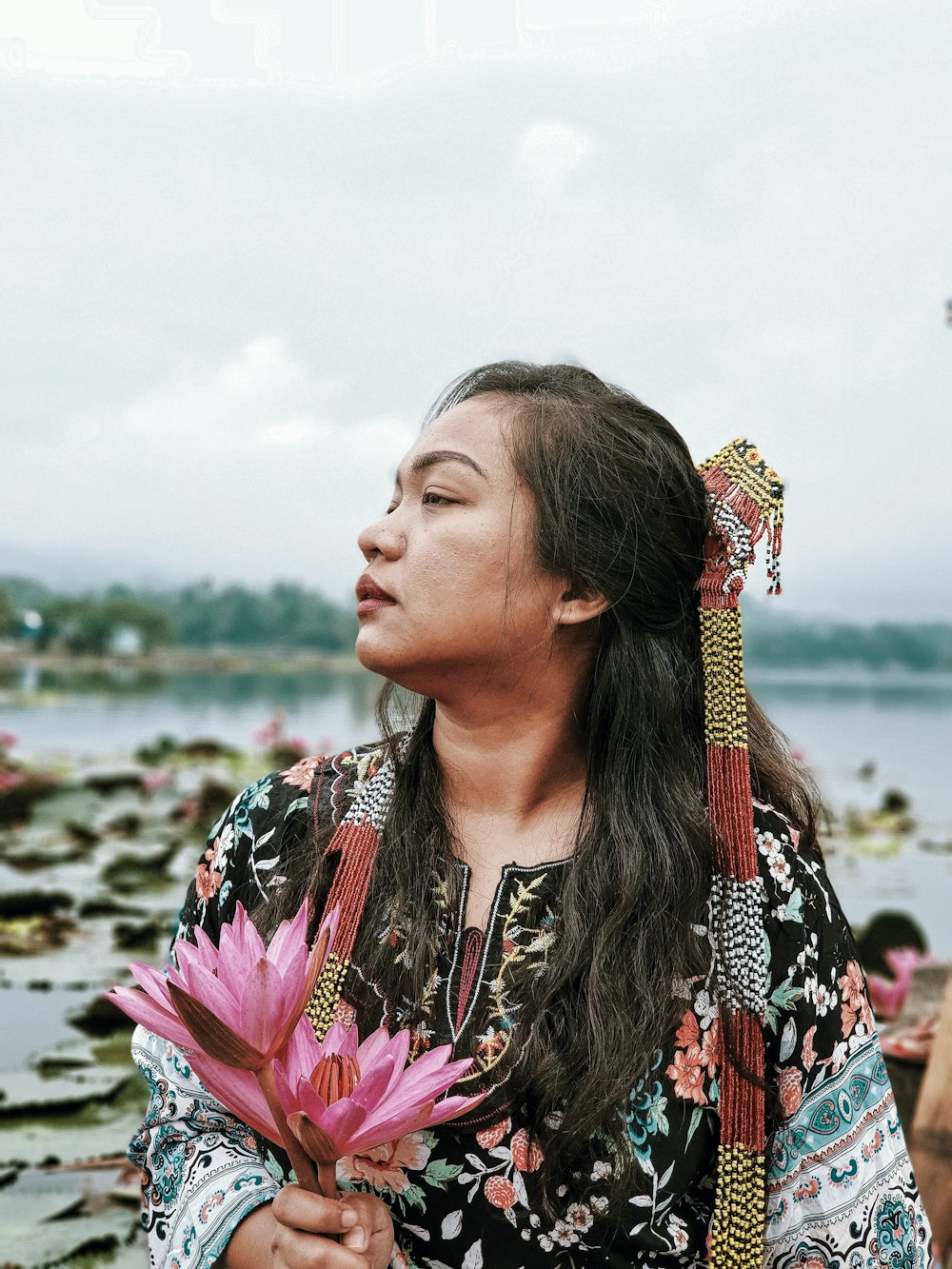 woman wearing black and multicolored floral long-sleeved blouse holding pink lotus flowers while facing her right right during daytime