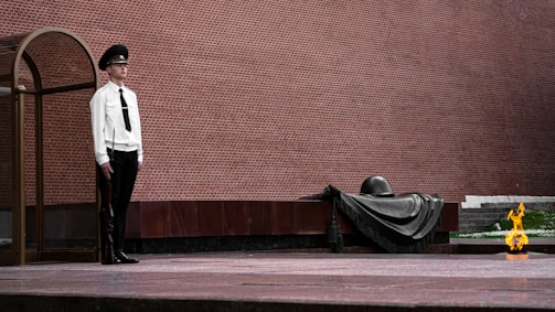 A guard in a formal uniform stands solemnly next to a glass enclosure, facing a monument. The monument features a shrouded helmet and a draped cloth, symbolizing remembrance. A small eternal flame burns nearby, adding a sense of reverence to the scene. The backdrop is a large, textured brick wall.