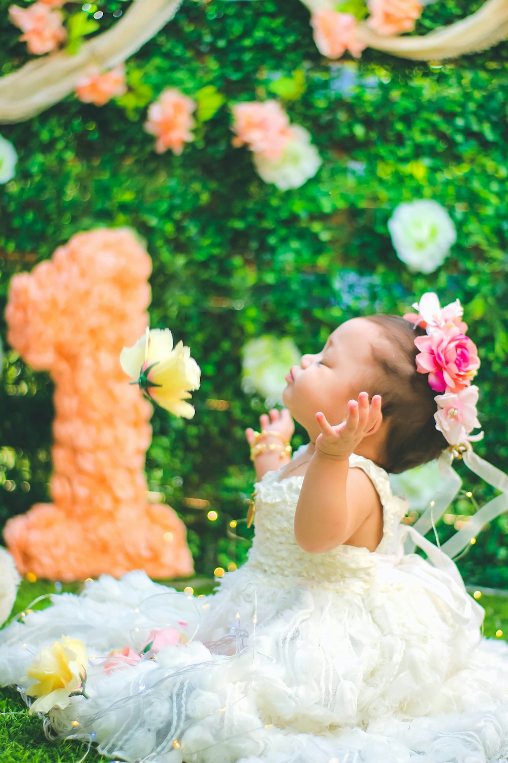 closed eyed baby girl wearing white gown sitting on grass and spreading her arms