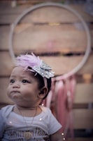 A baby is wearing a white shirt and a necklace with a heart pendant. A decorative headband with a lavender feather and silver bow adorns the head. The background features a large circular object with pink ribbons hanging underneath.