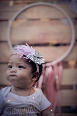 A baby is wearing a white shirt and a necklace with a heart pendant. A decorative headband with a lavender feather and silver bow adorns the head. The background features a large circular object with pink ribbons hanging underneath.
