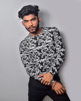 A person with neatly styled dark hair and facial hair is posing against a plain gray background. They are wearing a black and white patterned long-sleeve shirt and black pants, with a beaded bracelet on the wrist.