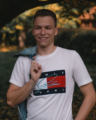A young person is smiling while holding a denim jacket over their shoulder. They are wearing a white T-shirt with a colorful graphic design. The background is blurred, featuring trees and foliage with sunlight filtering through.