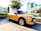 A bright yellow Mini Cooper with black racing stripes is parked on a patterned brick plaza. The car is positioned with a backdrop of modern buildings, lush greenery, a tall tree, and some white flowers. The surrounding area appears to be an urban setting with clear skies, suggesting daylight.