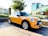 A bright yellow Mini Cooper with black racing stripes is parked on a patterned brick plaza. The car is positioned with a backdrop of modern buildings, lush greenery, a tall tree, and some white flowers. The surrounding area appears to be an urban setting with clear skies, suggesting daylight.