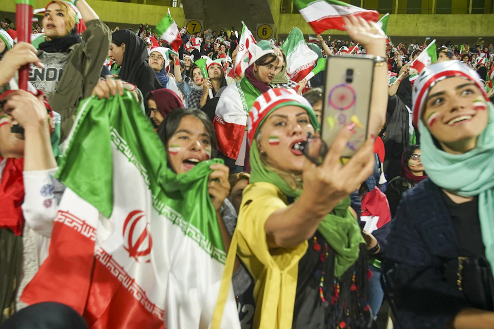 The image captures a lively scene at a sporting event, with a crowd of people gathered to cheer on their team. The majority of the spectators are wearing green and red clothing, which is consistent with the colors of the Iranian flag. Among them, there are two individuals who stand out - one person holding up an American flag, while another individual is capturing the moment by taking a photo using their phone.