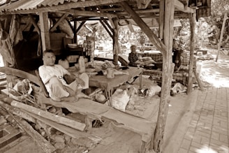 A rustic wooden shelter with three people seated inside offers a glimpse into a simple, rural life. The structure appears weathered, with a corrugated metal roof and wooden beams. Within the shelter, a man is sitting cross-legged on a wooden platform, while two older individuals are seated on a makeshift bench. Various objects and containers are scattered around, adding to the scene's lived-in feel. Trees and a clay-tiled path are visible in the background, suggesting an outdoor setting.