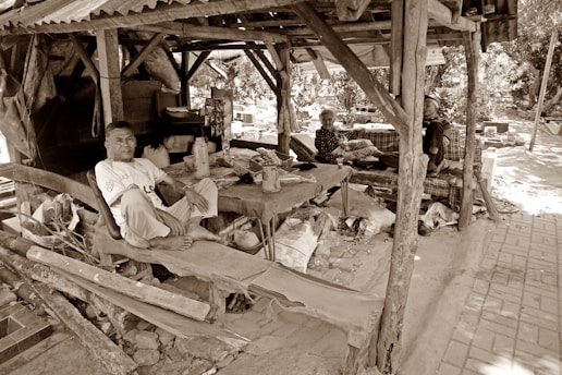 A rustic wooden shelter with three people seated inside offers a glimpse into a simple, rural life. The structure appears weathered, with a corrugated metal roof and wooden beams. Within the shelter, a man is sitting cross-legged on a wooden platform, while two older individuals are seated on a makeshift bench. Various objects and containers are scattered around, adding to the scene's lived-in feel. Trees and a clay-tiled path are visible in the background, suggesting an outdoor setting.