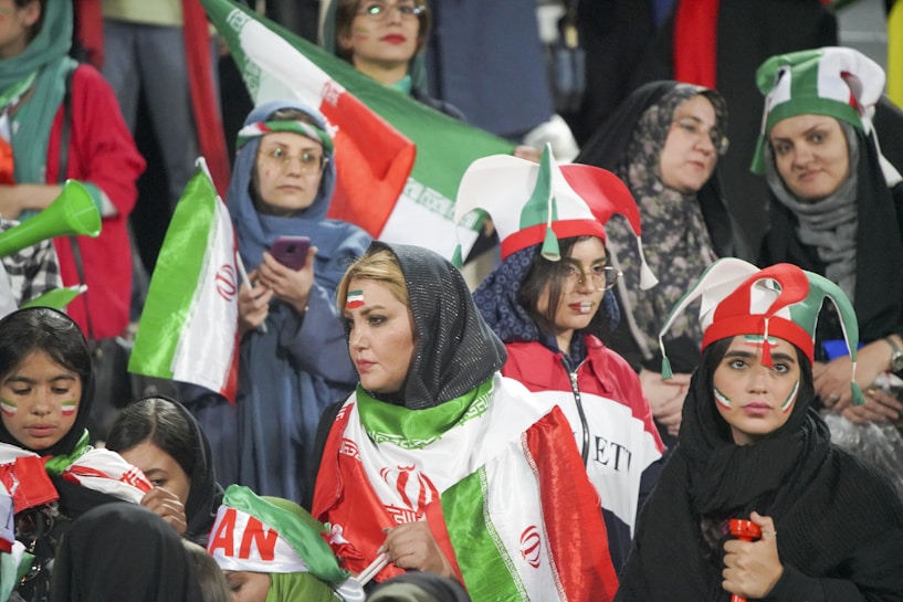 A group of women are dressed in colorful costumes and hold the Iranian flag. They have face paint and hats in red, white, and green, possibly showing enthusiastic support for their country at a sporting event.
