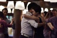 Couples are engaging in a close embrace dance in a warmly lit room, suggesting an intimate and focused atmosphere. The lighting is dim with soft overhead lights, creating a cozy setting. The individuals appear to be dancing in pairs, with attention to their partners.