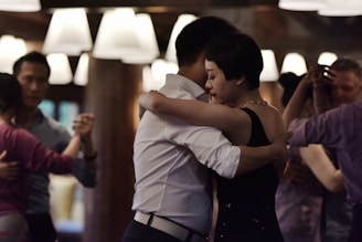 Couples are engaging in a close embrace dance in a warmly lit room, suggesting an intimate and focused atmosphere. The lighting is dim with soft overhead lights, creating a cozy setting. The individuals appear to be dancing in pairs, with attention to their partners.