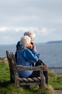 An elderly couple sits on a wooden bench overlooking a calm sea, with grassy surroundings. The woman is wearing a blue jacket, and the man is dressed in brown. Both have white hair and are relaxing while looking at the water.