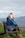An elderly couple sits on a wooden bench overlooking a calm sea, with grassy surroundings. The woman is wearing a blue jacket, and the man is dressed in brown. Both have white hair and are relaxing while looking at the water.