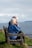 An elderly couple sits on a wooden bench overlooking a calm sea, with grassy surroundings. The woman is wearing a blue jacket, and the man is dressed in brown. Both have white hair and are relaxing while looking at the water.
