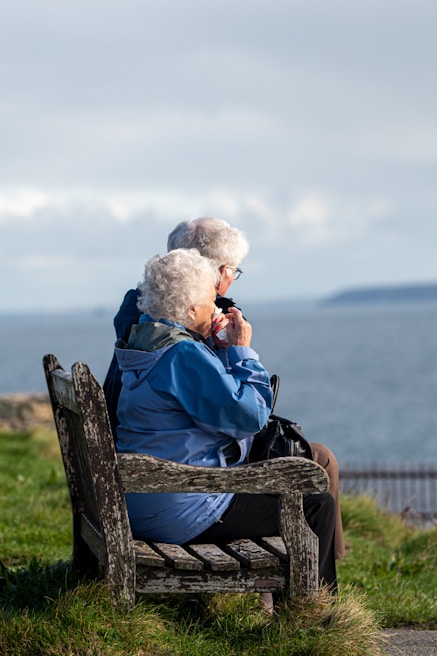 An elderly couple sits on a wooden bench overlooking a calm sea, with grassy surroundings. The woman is wearing a blue jacket, and the man is dressed in brown. Both have white hair and are relaxing while looking at the water.