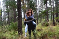 A young couple enjoys a playful moment in a dense, green forest. The man carries the woman on his back as they smile and share a joyful interaction. Surrounding them are tall pine trees and lush greenery, creating a serene and natural setting.