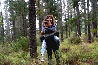A young couple enjoys a playful moment in a dense, green forest. The man carries the woman on his back as they smile and share a joyful interaction. Surrounding them are tall pine trees and lush greenery, creating a serene and natural setting.