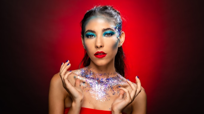 A woman with striking makeup featuring bright blue eyeshadow and bold red lipstick. Her face and shoulders are adorned with sparkling glitter, creating a dramatic and artistic look. The background is a gradient of red to brown, enhancing the vibrant colors of her makeup and the glitter.