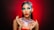 A woman with striking makeup featuring bright blue eyeshadow and bold red lipstick. Her face and shoulders are adorned with sparkling glitter, creating a dramatic and artistic look. The background is a gradient of red to brown, enhancing the vibrant colors of her makeup and the glitter.