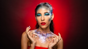 A woman with striking makeup featuring bright blue eyeshadow and bold red lipstick. Her face and shoulders are adorned with sparkling glitter, creating a dramatic and artistic look. The background is a gradient of red to brown, enhancing the vibrant colors of her makeup and the glitter.