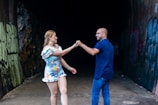 A man and a woman are dancing together in a dimly lit tunnel. The woman is wearing a colorful floral dress, and the man is dressed in a dark blue shirt and jeans. Graffiti is visible on the walls of the tunnel, adding an urban feel to the scene.