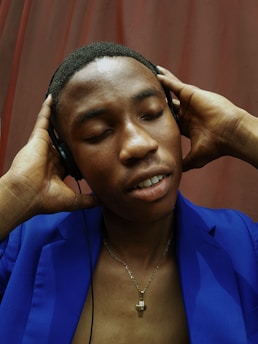 A person is wearing headphones with a relaxed expression, eyes closed, enjoying music. The individual is dressed in a blue blazer and a necklace, with their hands resting on the headphones. A brownish curtain serves as the background, adding a warm contrast to the scene.