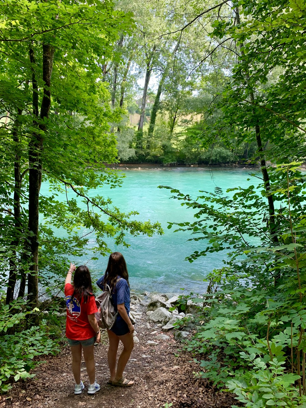 two women standing beside the river under tall trees