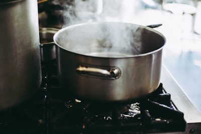 A stainless steel pot boiling with steam rising on a gas stove in a kitchen setting. The pot is prominently placed, while the background features blurred kitchen elements.